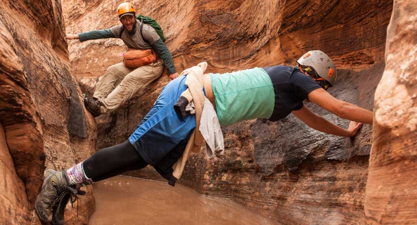 Two people wearing helmets brace themselves on either side of a slot canyon using their arms and legs. 